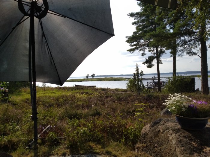 Solparasoll vid en sjöutsikt i en trädgård, omgiven av grönska och blommor, med brygga och klar himmel i bakgrunden.