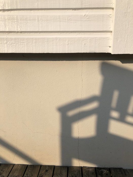Nymålad husgrund i beige med små sprickor, skugga av staket på trätrall.