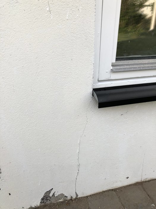 Sprickor i målad grundvägg av hus nära ett fönster, med tecken på slitage och avflagning.