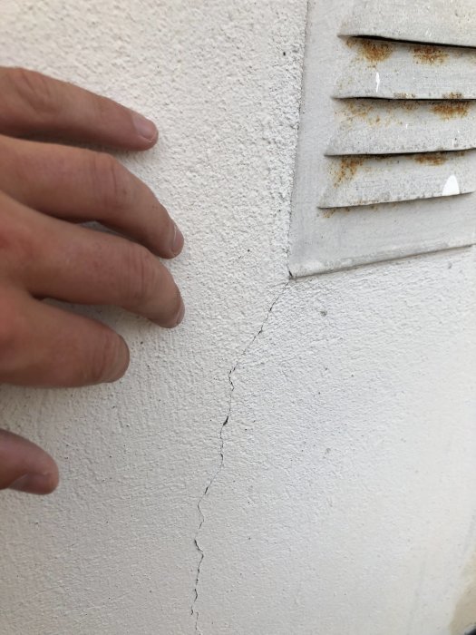 Närgränsad bild av en hand som pekar på en liten spricka i en målad husgrund nära en ventilspröjs.