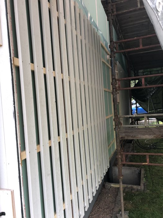 Vertikala träpaneler delvis monterade på ett hus med byggställningar runt omkring.