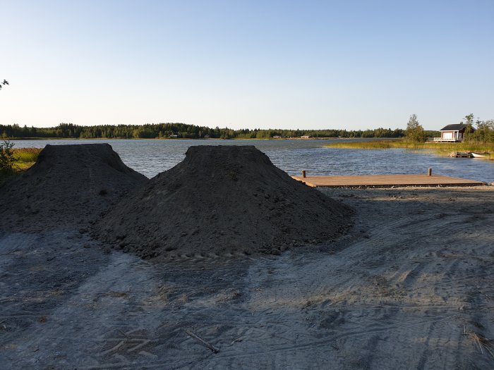 Högar av matjord levererade vid sjö med brygga och fritidshus i bakgrunden.