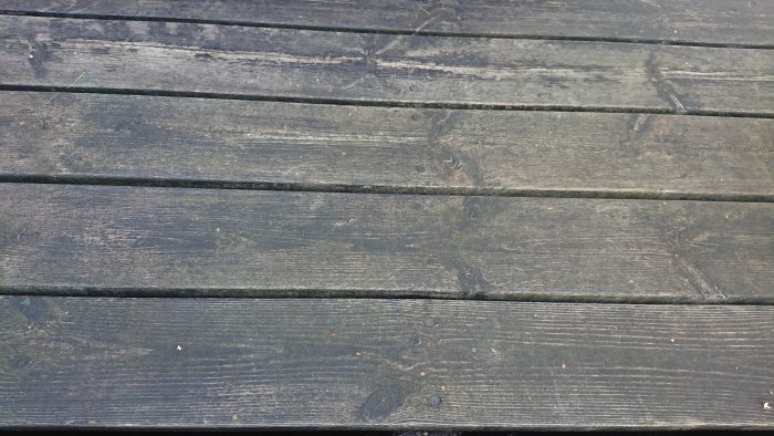Sliten svartlaserad veranda med synlig mossa och alger mellan plankorna som behöver underhåll.