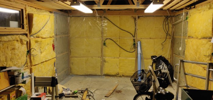 Oisolerat garage under renovering med isoleringsmaterial på väggarna och lösa elledningar som hänger framför.