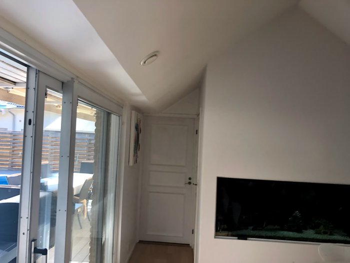 Interiör av ett hus med vitmålade väggar och lutande tak, glasdörrar till en terrass och ett akvarium inbyggt i väggen.