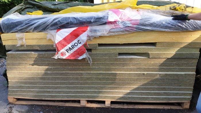 Stapel av gula klimatskivor på en träpall delvis täckt av en genomskinlig plast med märkningen PAROC.