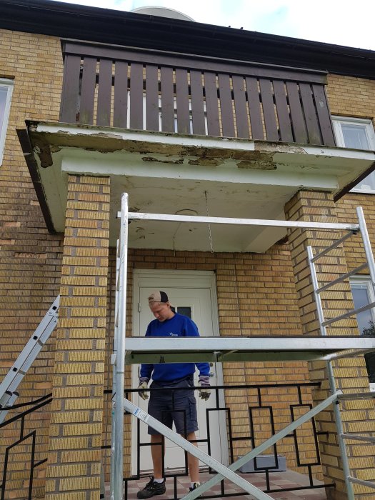 Person på byggnadsställning under en skadad balkonggolv med flagnande färg och synlig betong.