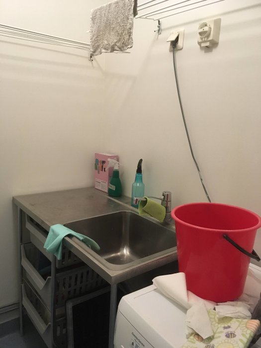 Tvättstuga med tvättmaskin, rostfri bänk med diskho, röd hink och tvättmedel, samt tvättmaskinsbrytare på väggen.