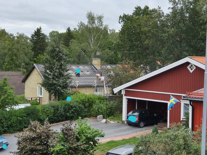 Vy över ett bostadsområde med gult hus, rött garage med öppna dörrar och Svenska flaggan, fråga om bygglov.