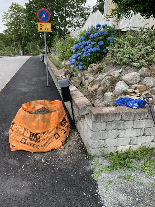 En lutande mur längs en väg med en stor säck grus och utrustning för reparation.