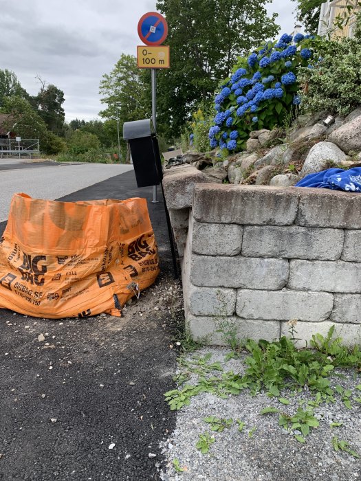 Mur av gråa betongblock som lutar framåt nära en väg med ett skadedepåsäck och blå hortensiabuskar i bakgrunden.