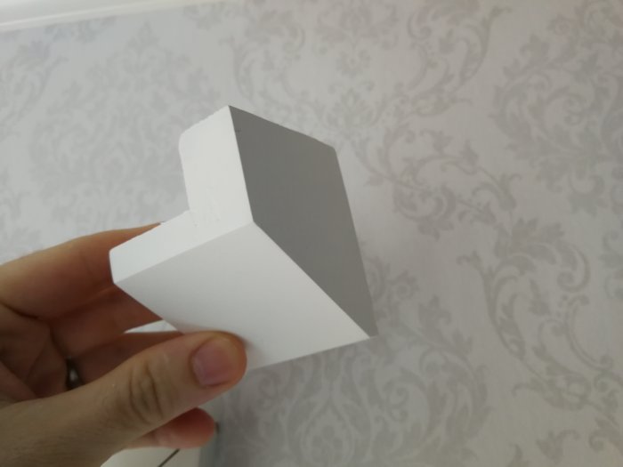 Hand håller en hemgjord vit kloss av trä mot en tapetserad vägg, klossen liknar en Legobit.