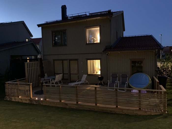 Ett modernt hus med ett nybyggt trädäck och horisontella räcken i skymningen, inklusive en blomlåda och utemöbler.