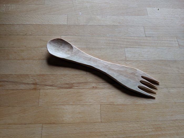 Handgjord kombinerad sked och gaffel i trä, kallad spork, på ett trägolv.