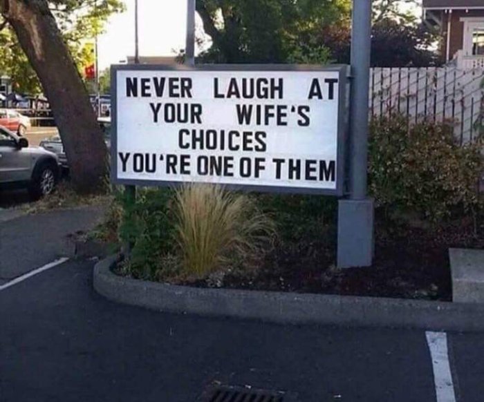 Skylt med texten "NEVER LAUGH AT YOUR WIFE'S CHOICES YOU'RE ONE OF THEM" vid en parkering.