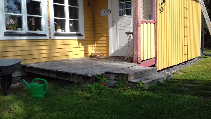 Gammal sliten träfarstukvist i behov av renovering utanför en gul husvägg med vit dörr och fönster.