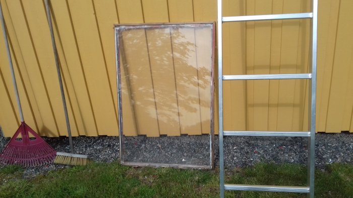 Demonterat plexiglasfönster lutat mot en gul vägg bredvid en stående stege, med en röd kratta och gul sopborste på marken.