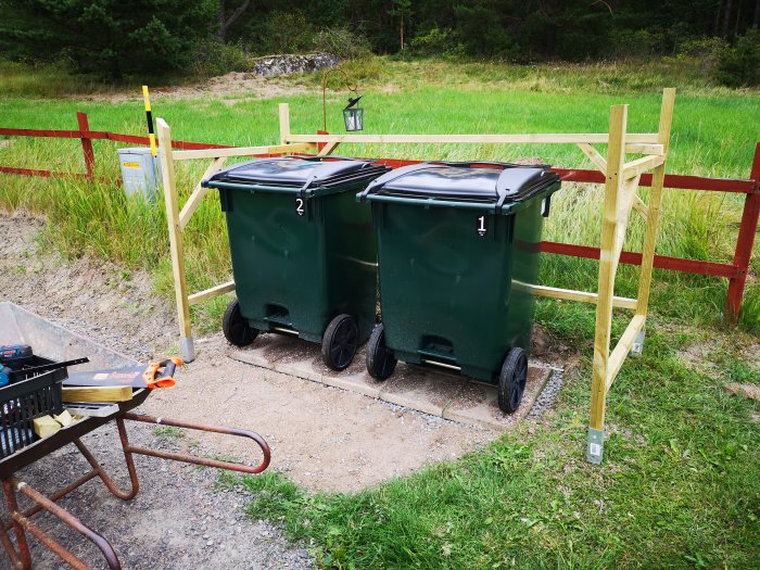 Pågående bygge av ett trähägn för soptunnor, verktyg inklusive skottkärra syns i förgrunden.