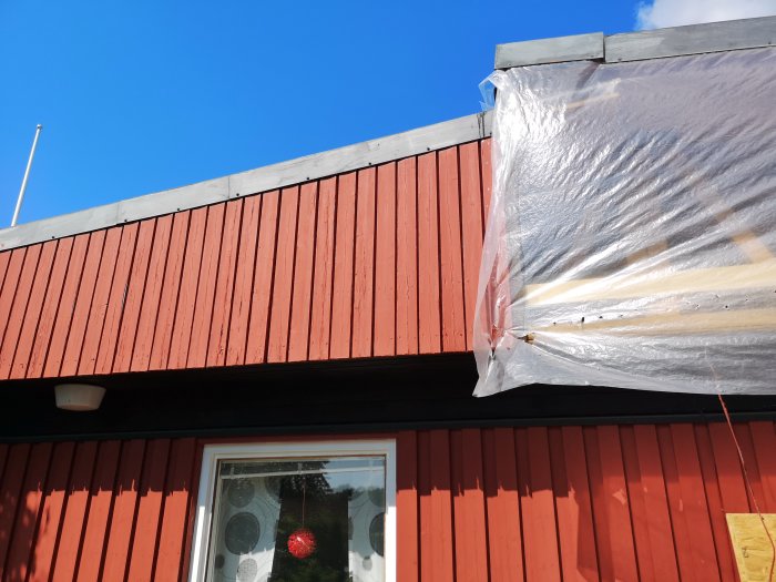 Renoverat hus med röd träpanel och synlig asfaboard bakom, delvis täckt med plast.