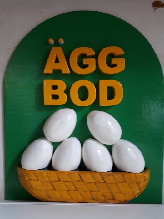 Tredimensionell skylt med texten "ÄGG BOD" i gula bokstäver och illustration av vita ägg i en gul korg mot grön bakgrund.