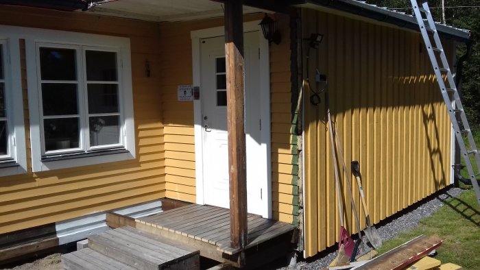 Delvis renoverat gult hus med ny grundmålad panel och oljebehandlade reglar intill en trätrall och en stege.