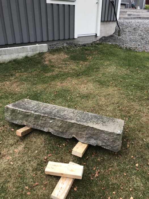 Stor bohuslänsk granitsten avsedd som trappsteg, upplyft på träklossar på en gräsmatta framför en byggnad.