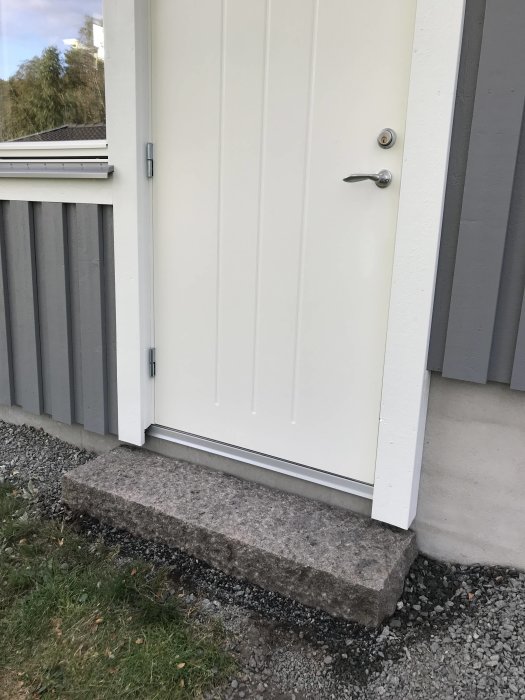 Stor granitsten använd som trappsteg framför dörr till vit snickarbod, omgivet av grus och gräs.