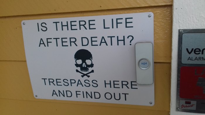 Förmanande skylt med texten "IS THERE LIFE AFTER DEATH? TRESPASS HERE AND FIND OUT" bredvid en ringklocka.
