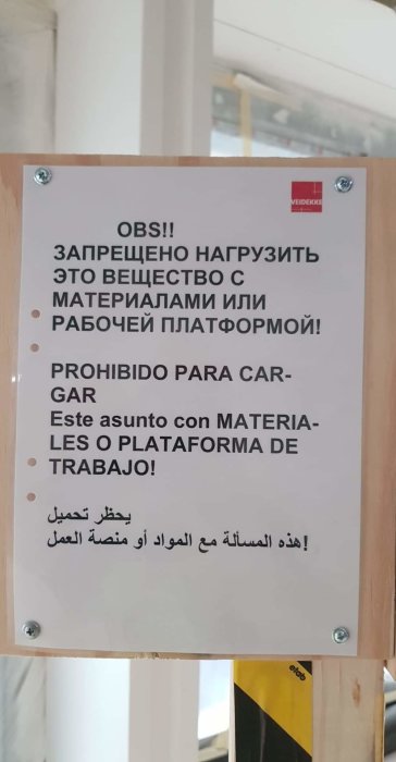 Varningskylt på flera språk fäst på en trästomme med säkerhetsinstruktioner på byggarbetsplats.