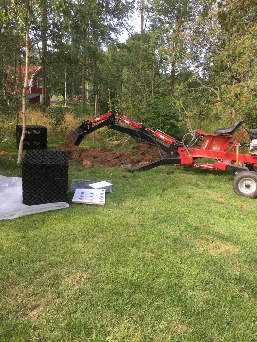 Minigrävare i arbete med att gräva ner stenkistor till dagvatten på en gräsmatta med hus och träd i bakgrunden.