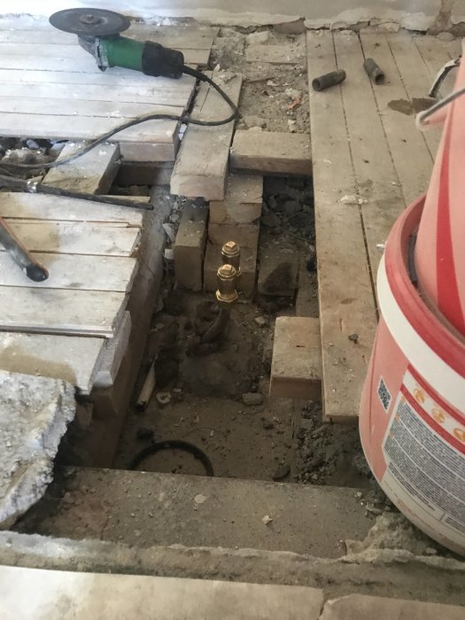 Öppnad golvsektion med rör och blandade plankgolv, verktyg och skyddspapp vid renovering.