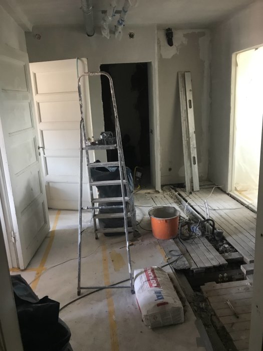 Renoveringsarbete i en hall med en stege, olika golvtyper och ett öppet golv där nya rör installerats.