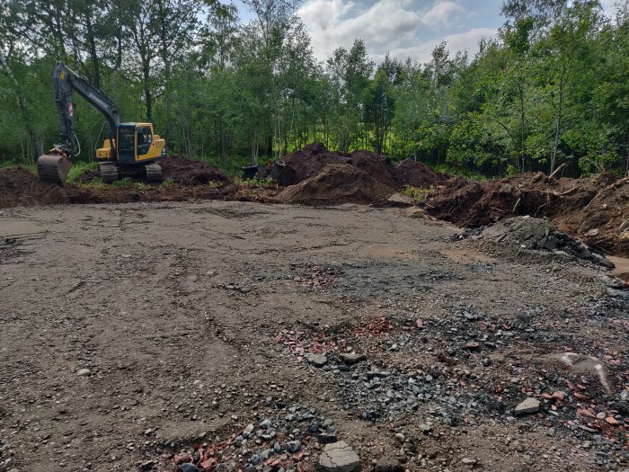 Grävare på en tom byggplats som har tagit bort jord och förbereder för fyllnadsmassor i en skogsmiljö.