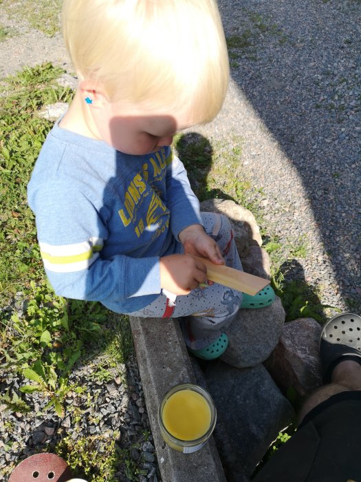 Liten pojke som hjälper till med att slipa träbit utomhus bredvid en vuxen och ett glas med vätska.