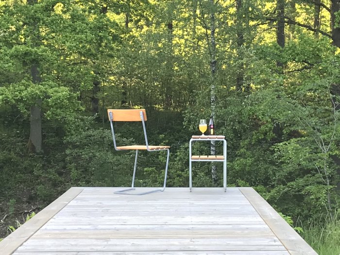En brygga med en ensam stol och ett litet bord med en flaska och två glas, omgiven av gröna träd.