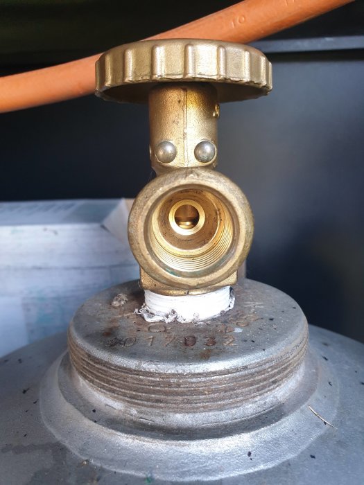 Mässingsventil fäst på en stor metallbehållare med tejpade gängor, mot mörk bakgrund.