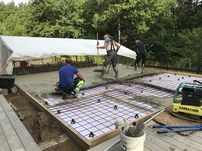 Arbetare häller betong på armeringsnät för grundläggning under ett vitt tält med skog i bakgrunden.