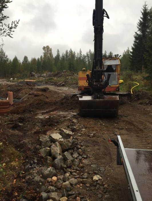 Grävmaskin på skogstomt som arbetar med markberedning, omgiven av sten och lera.