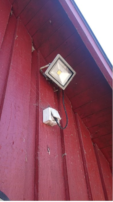 Strålkastare monterad på röd vägg med gummikabel till kopplingsdosa, synliga sockerbitar inne i dosan.