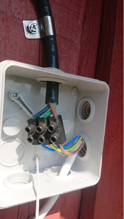 Eluttagsdosa med synliga ledningar och en "sockerbit" för elkopplingar mot en röd vägg, indikerar en amatörmässig installation.