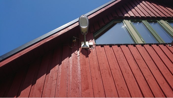 En strålkastare monterad på en röd trävägg under ett hustak med synlig elinstallation och kabeldragning.
