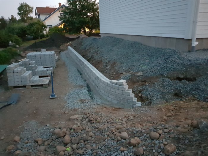 Nyanlagd stödmur och grus vid hus i skymningen, med skottkärra och stenblock i förgrunden.