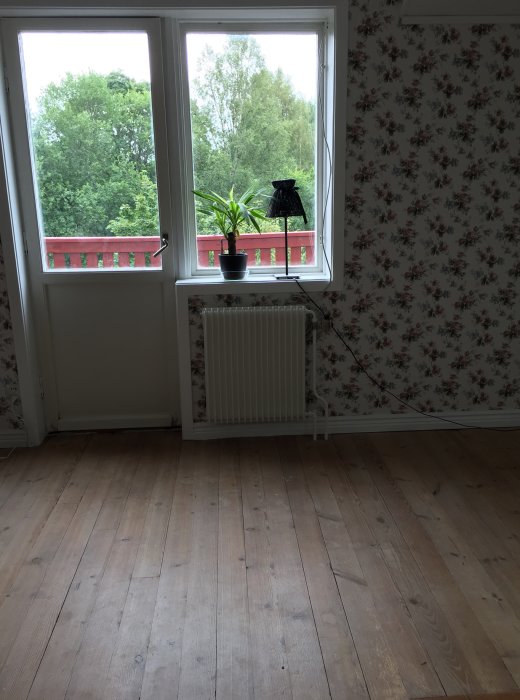 Bild av ett rum med slitet trägolv, vita fönster med utsikt, element och blommigt tapetvägg.