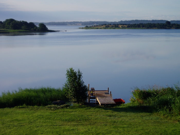 Vy över stilla vatten från Södra Fäjö med brygga och röd båt, med ö Senoren i bakgrunden.