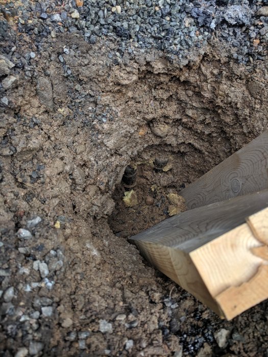 Grävd grop med jord, sand, stenar och synlig gul isolering vid rörutgång under träplanka.