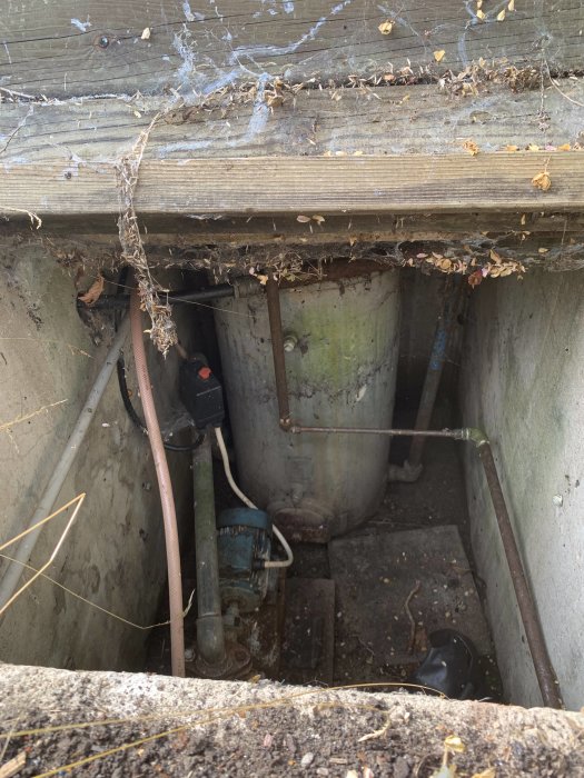 Öppet gjutet hål i mark med en äldre vattentank och vattenpump under trälock, omgiven av spindelnät och löv.