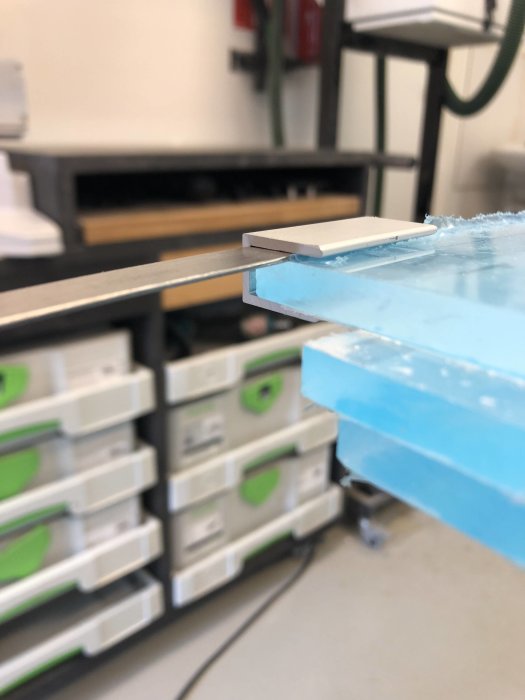 Närbild på en U-profil med 1 mm shim mellan profil och kantad plexiglasskiva, bakgrund med förvaringslådor.