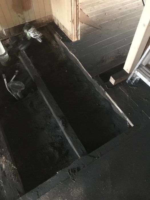 Renoveringsarbete med borttaget golv och synliga bjälkar täckta av kol och isolering i ett blivande badrum.