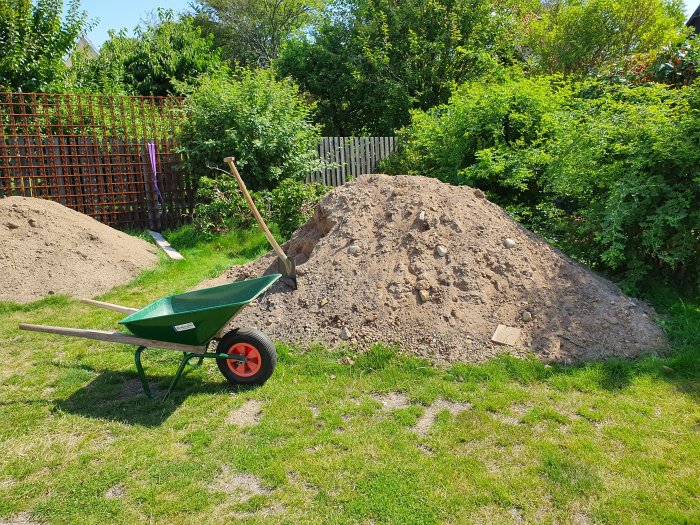 Grön skottkärra framför en stor grushög i en trädgård, arbetsstart för ett byggprojekt.