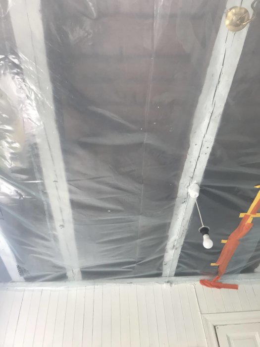 Oavslutat innertak med synliga träbjälkar och en påbörjad installation av diffusionspärr, tillsammans med en hängande glödlampa.